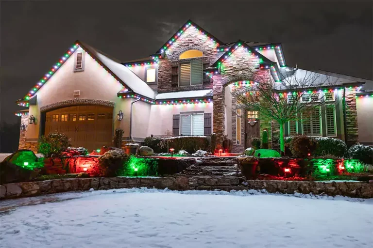 Luxury home exterior with Christmas holiday lighting featuring multicolored roof lights and red-green landscaping lights in snowy winter setting.