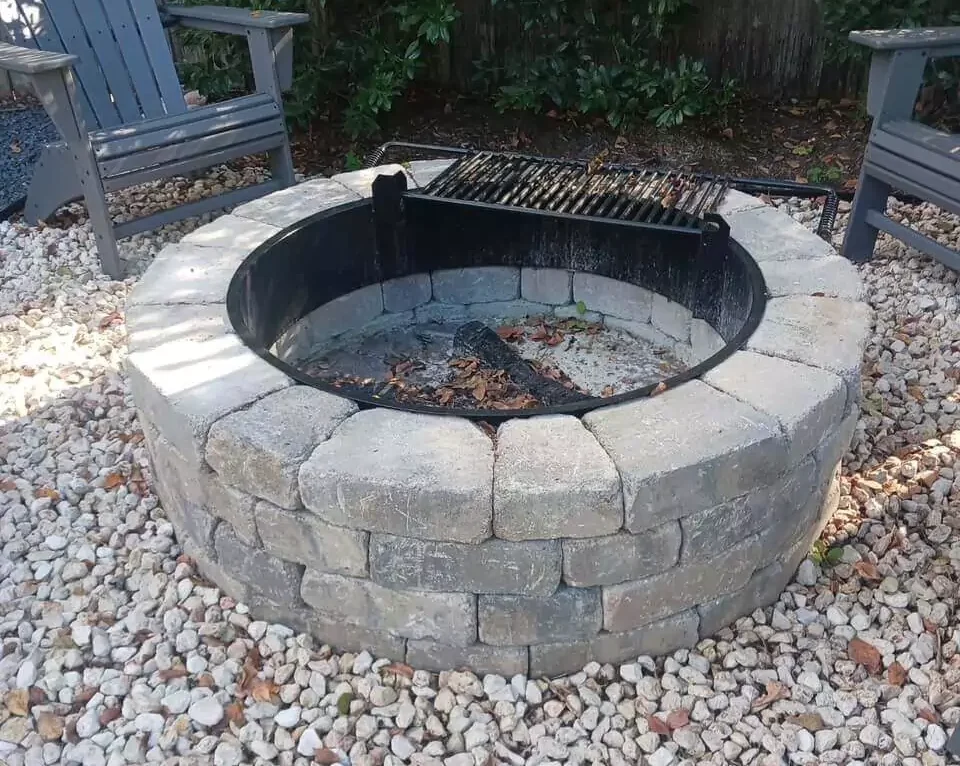 Custom stone fire pit with built-in grill surrounded by gravel and Adirondack chairs – perfect for outdoor gatherings. Expertly designed by Pearce Scapes.