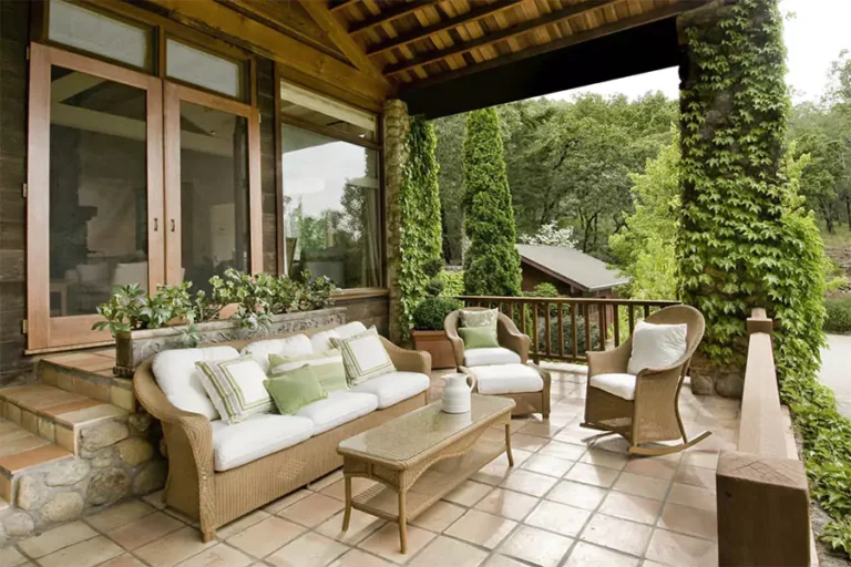 Cozy outdoor patio with wicker furniture, green cushions, stone tiles, and lush greenery.