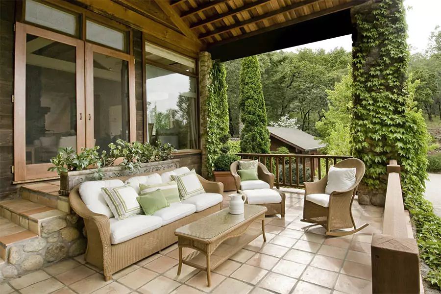 Cozy outdoor patio with wicker furniture, green cushions, stone tiles, and lush greenery.