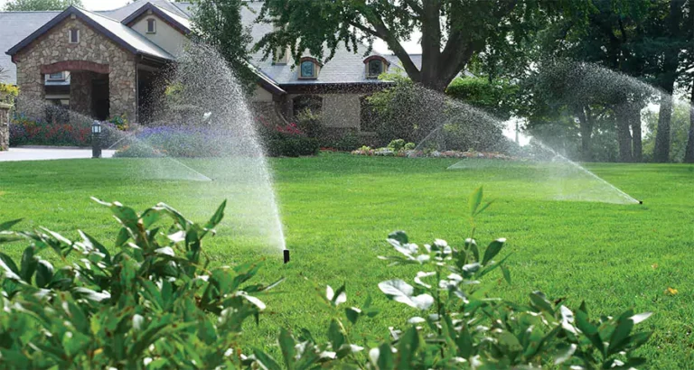 Automatic sprinkler system watering lush green lawn in front of residential home with flower beds
